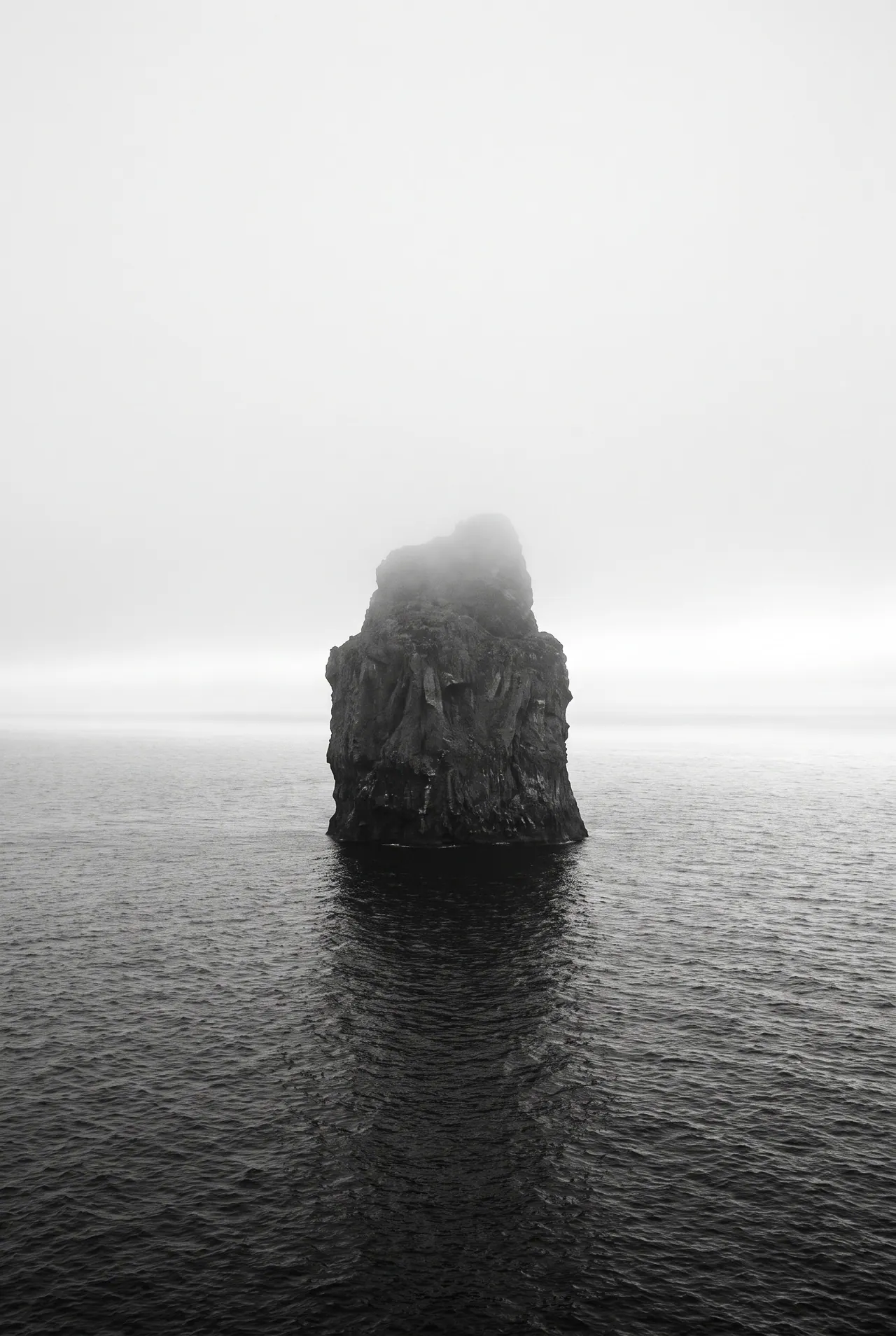 In black and white, a tall sea stack rises out of dark water, its summit dissolved in cloud, the sea around it textured with fine wind ripples.