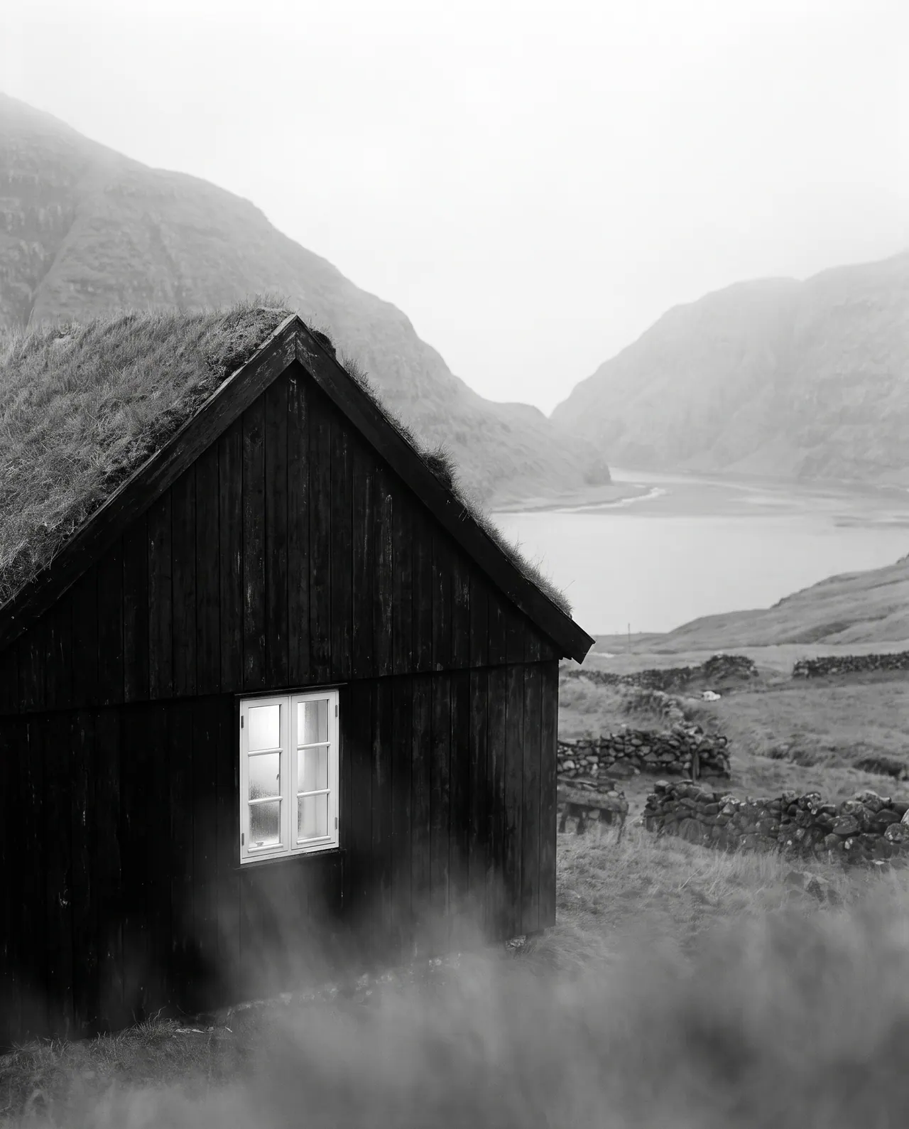 In black and white, a close view of a dark timber house with a grass roof. Its single white-framed window is the brightest point in a soft grey scene.