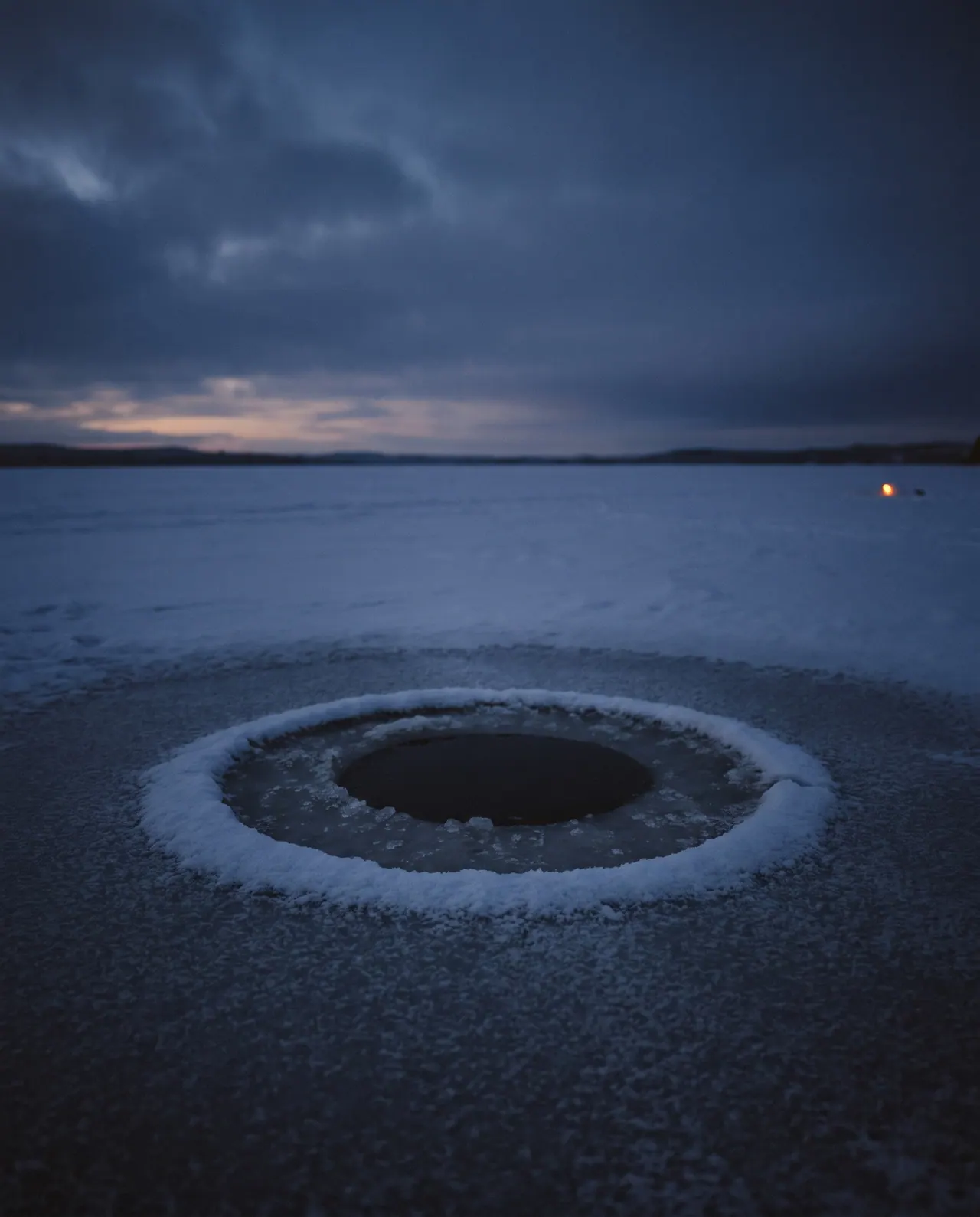 A round hole cut through thick lake ice, dark water inside it and a thin rim of fresh snow around its edge.