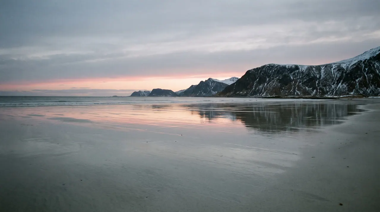 A wide, empty beach at blue hour. Wet sand mirrors a pale sky, and a dark mountain ridge sits low on the horizon beneath a faint band of rose-coloured light.