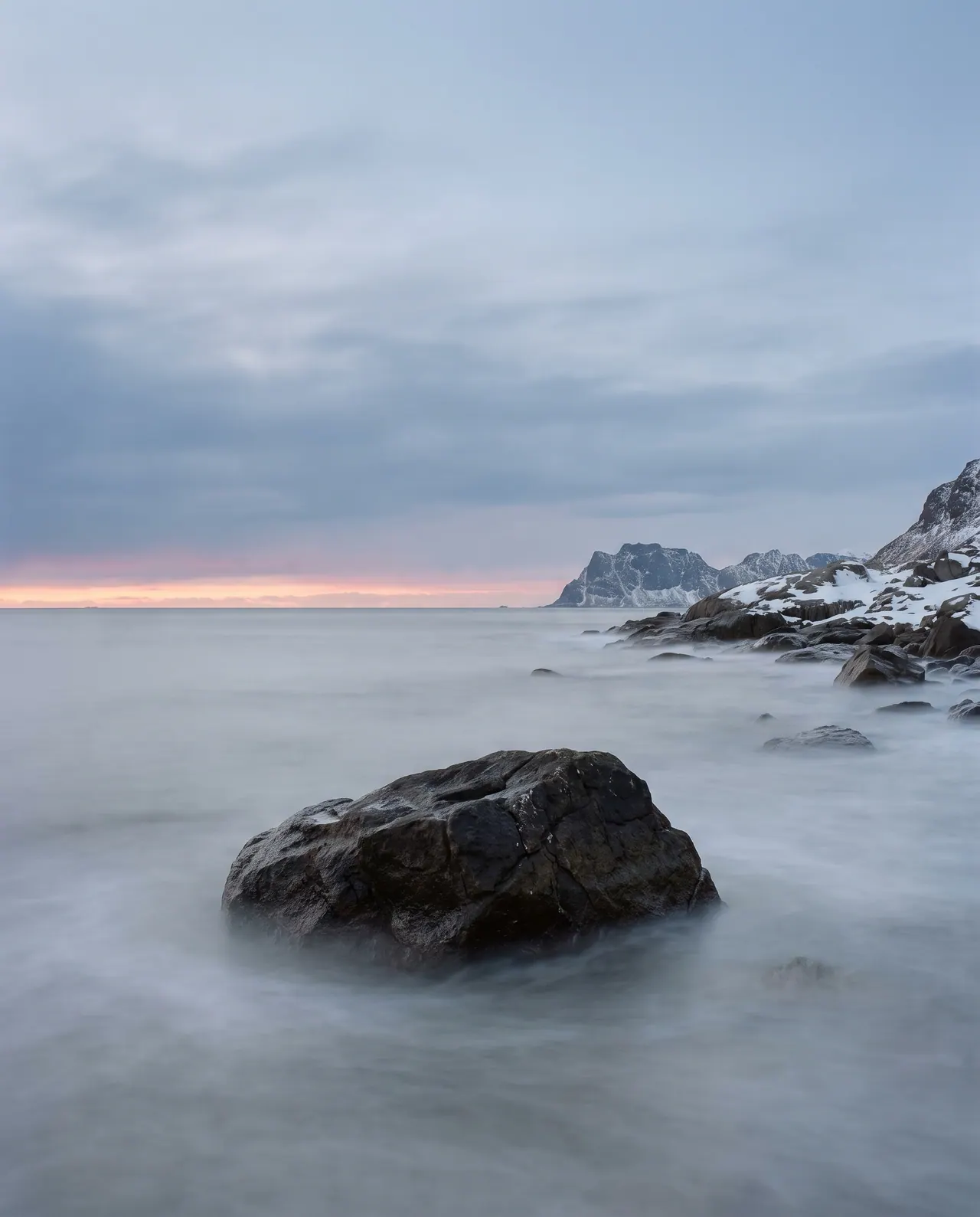 A long exposure turns the sea into a smooth silver mist around a single dark boulder in the foreground. The horizon is barely visible.